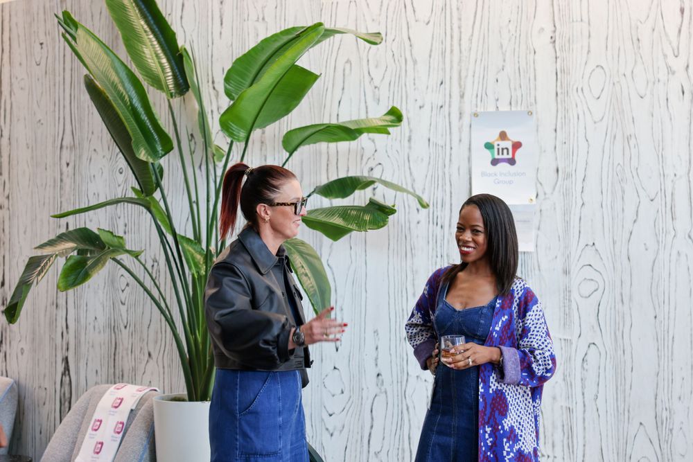 Two women smiling and chatting near a large tropical plant against a wood-paneled wall at a casual event.