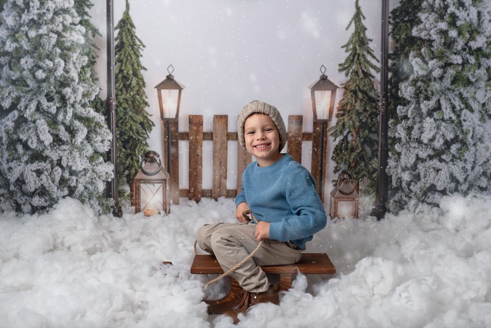 A winter wonderland photo setup with snowy evergreen trees, wooden fence, lanterns and white fluffy snow on the ground.