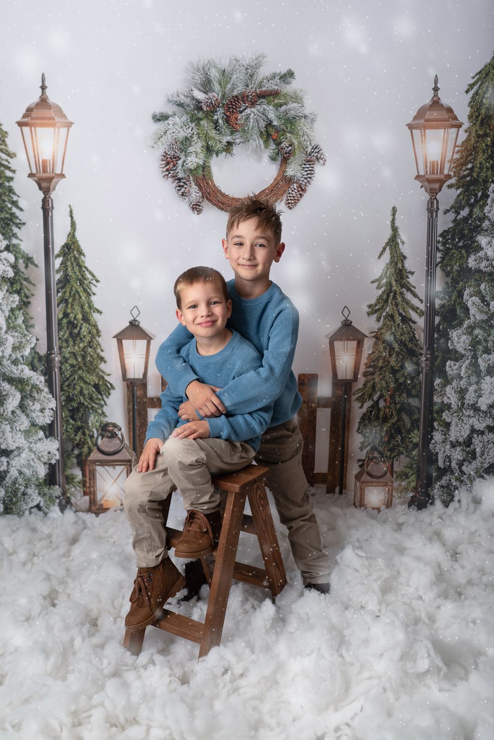 Winter holiday photo session with snowy scene backdrop featuring copper lanterns, evergreen branches and wooden chair decor.
