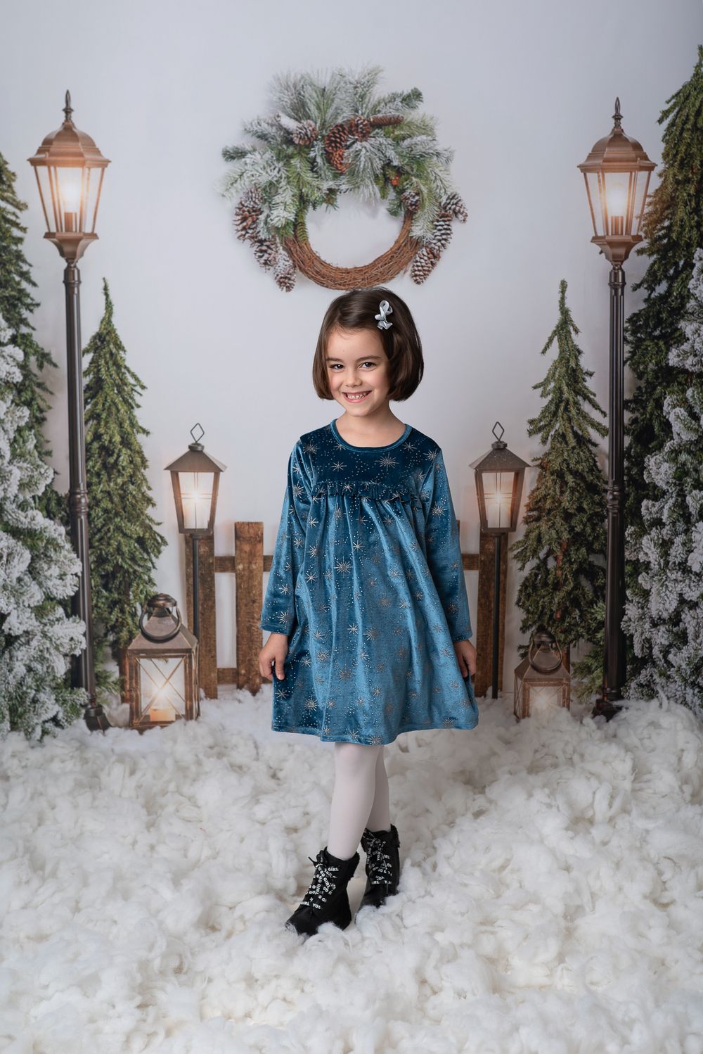 A winter photo session featuring a person in a teal velvet dress against a snowy backdrop with lanterns and evergreens.
