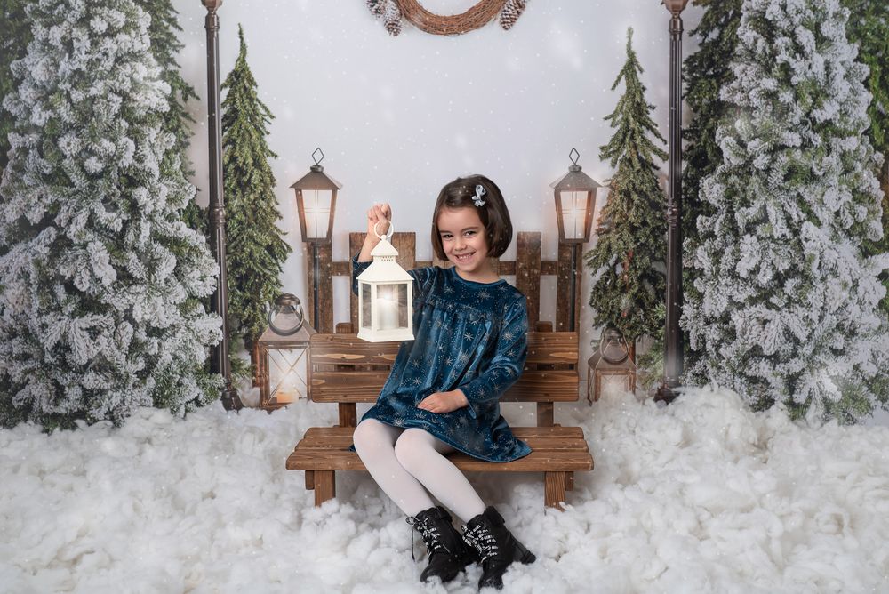 A winter wonderland photo session setup with rustic wooden bench, lanterns and artificial snow against a white backdrop.