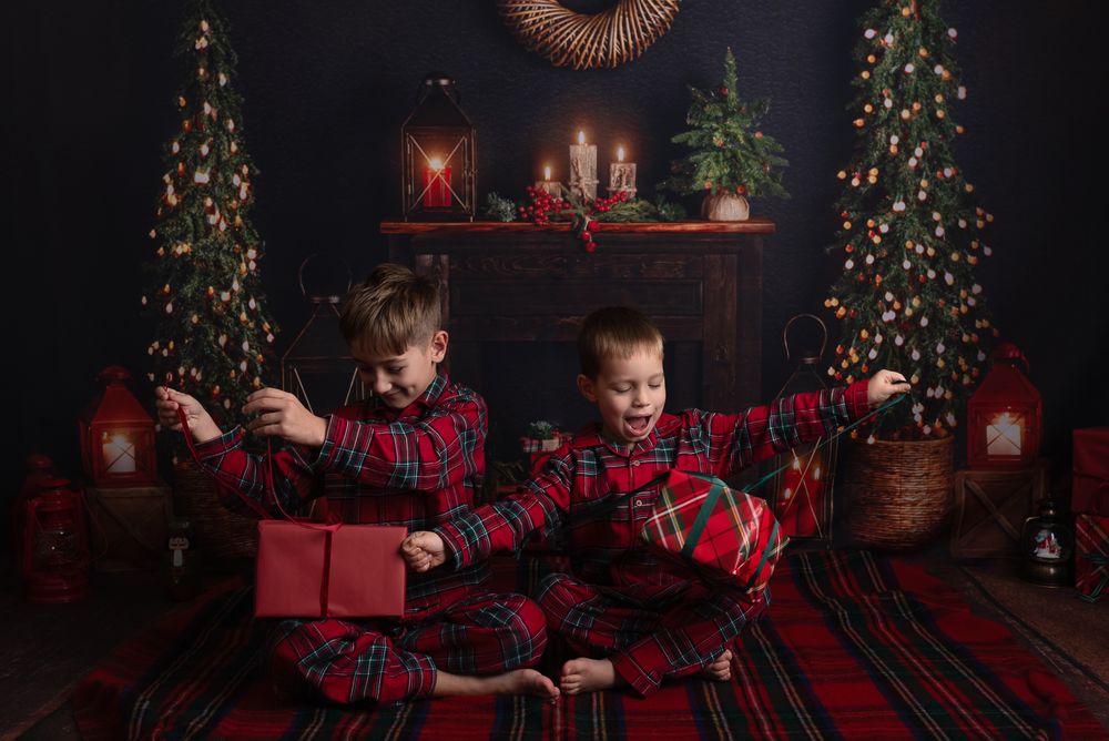 Children in matching buffalo plaid outfits open Christmas presents in front of decorated trees in a dark studio setting.