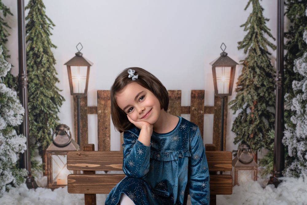 Woman in denim jacket poses against wooden fence with lanterns and evergreen trees in winter setting.