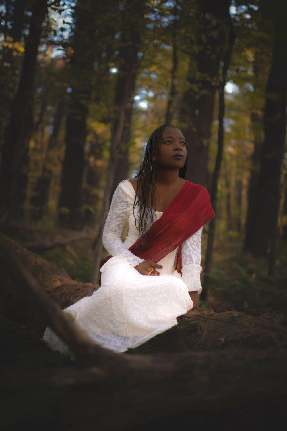 Figure in white dress with red sash sits thoughtfully in autumn forest.