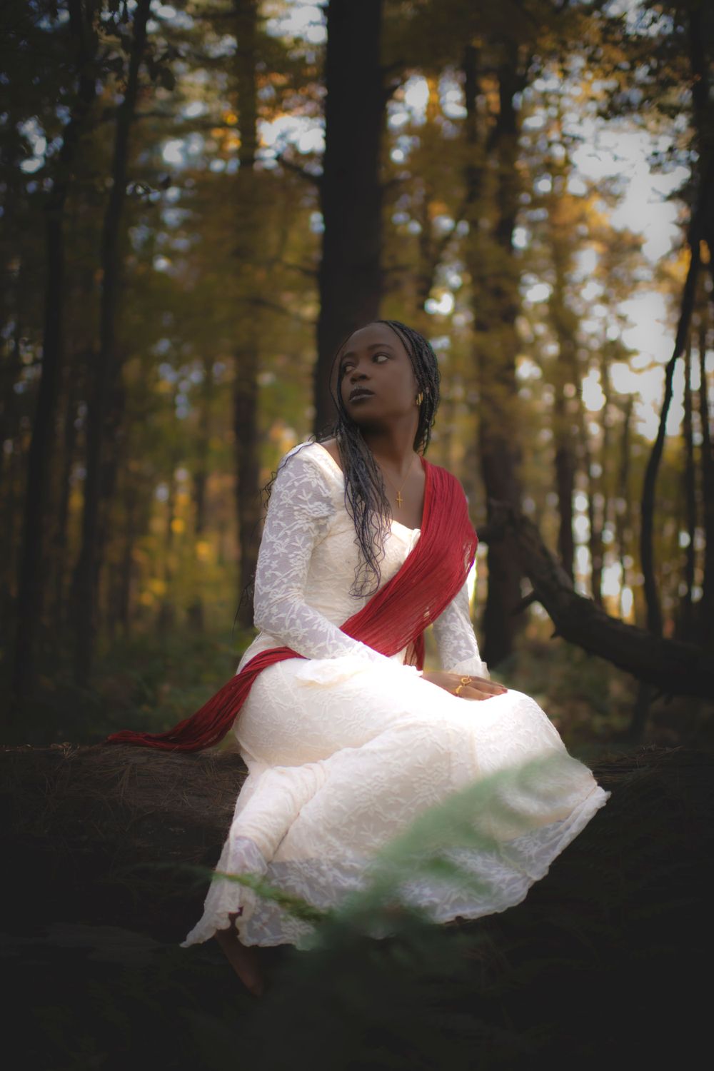 Contemplative pose in white dress and red sash amidst golden forest setting.