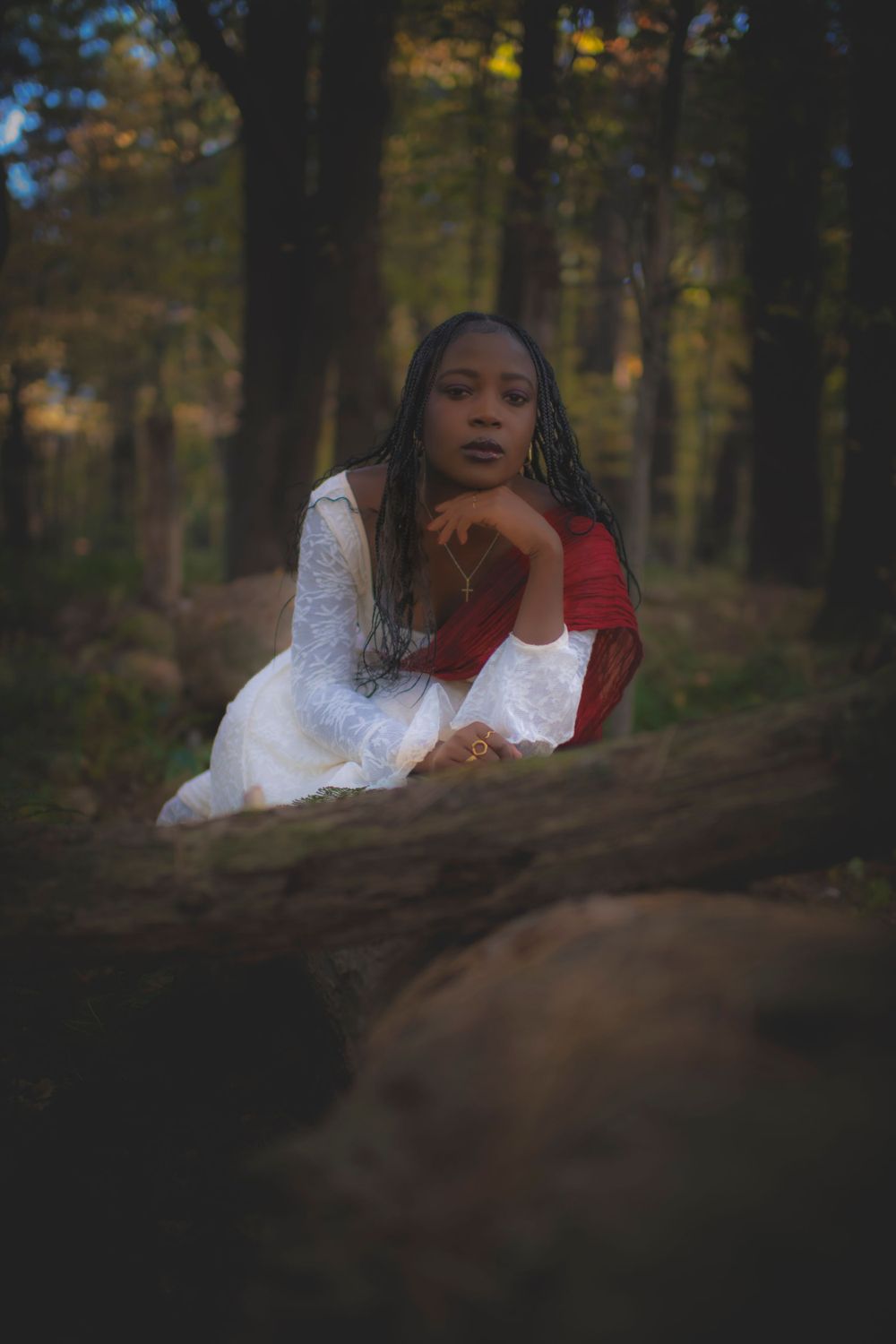 Ethereal portrait in a white dress and red sash resting thoughtfully against autumn forest backdrop.