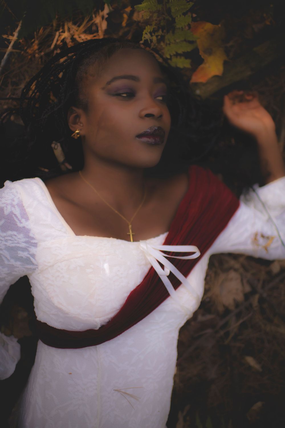 Artistic portrait featuring white lace dress with burgundy sash against dark forest background.