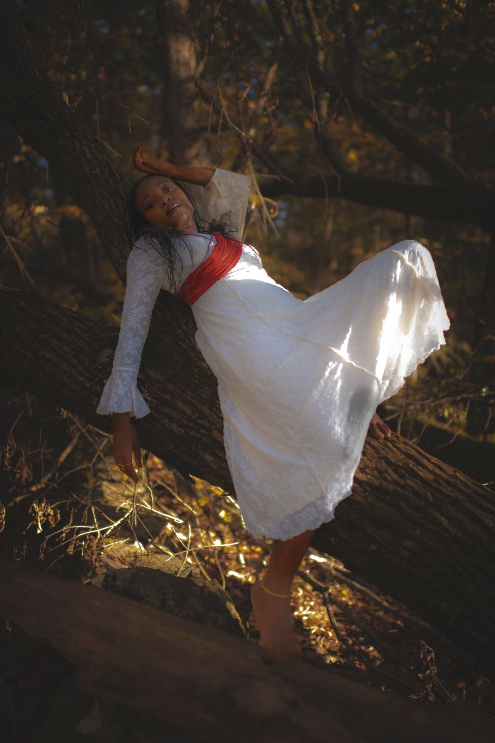 Dynamic pose in white vintage dress with red sash against golden forest light.