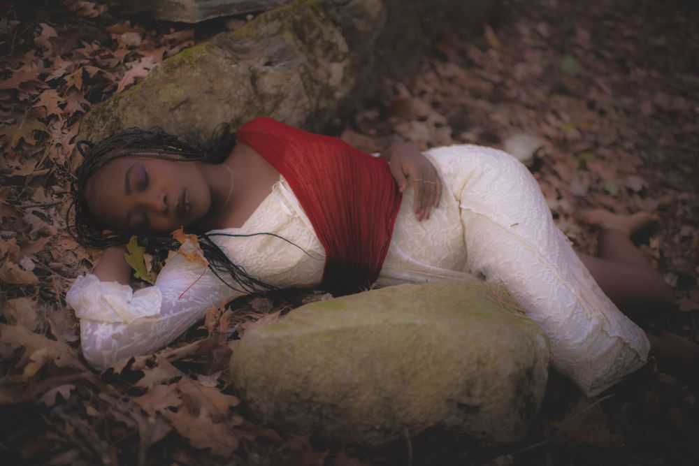 Artistic portrait on forest floor with white dress and red wrap against natural stone.