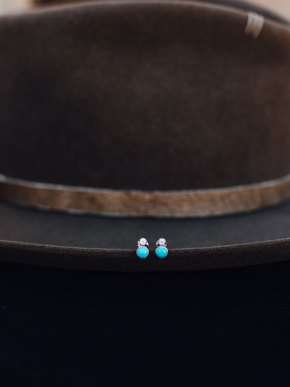 Tiny turquoise stud earrings displayed on brim of dark brown hat.