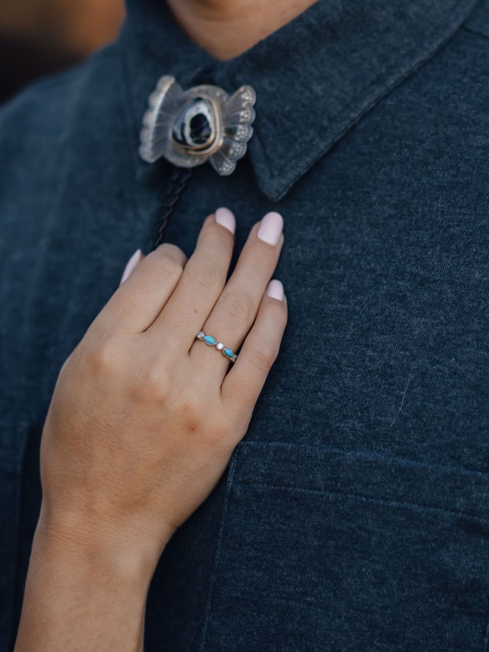 A hand wearing a delicate ring touches a dark denim collar with ornate brooch.