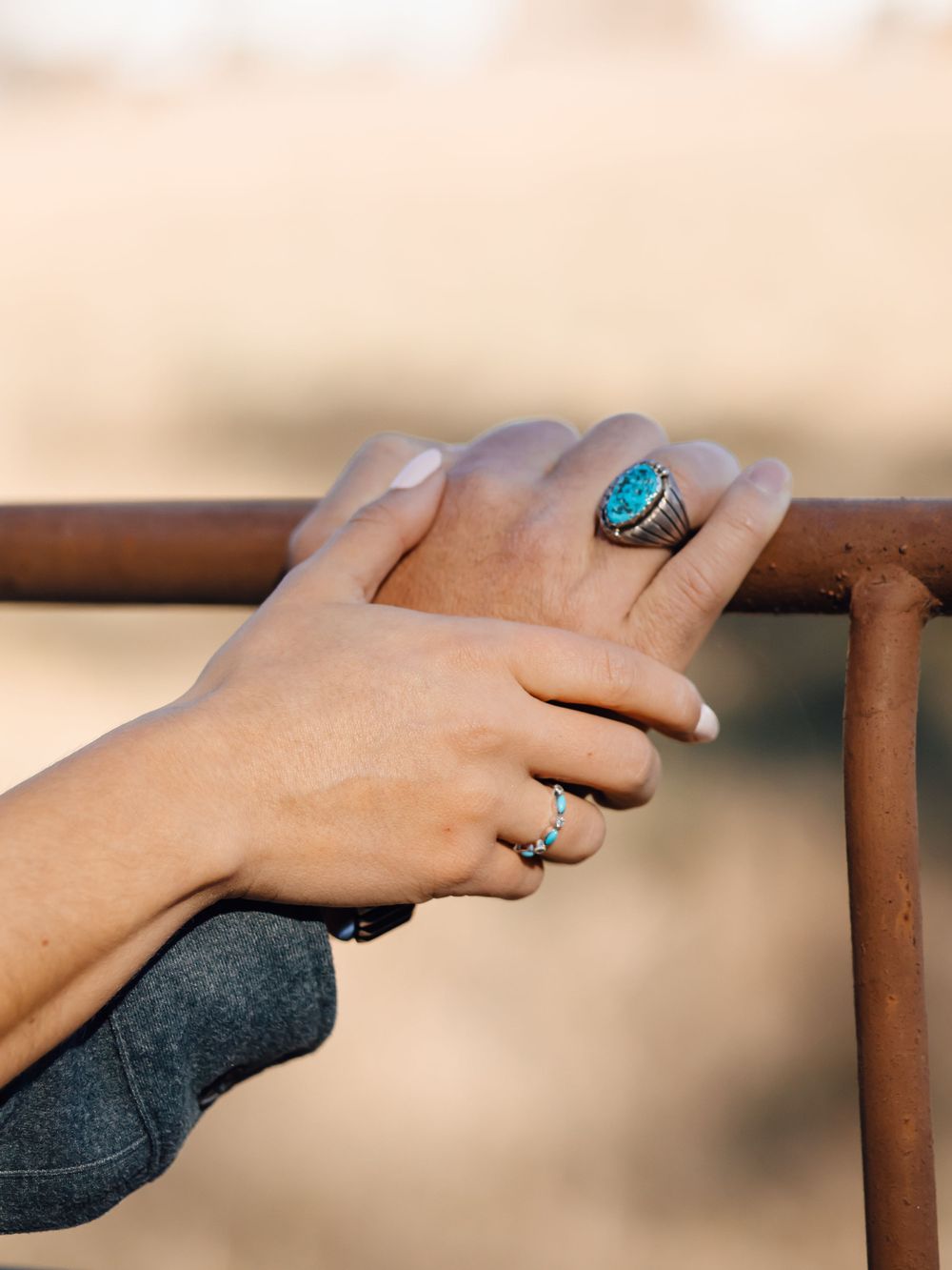 Hand wearing turquoise statement ring and delicate band resting on rustic metal railing.