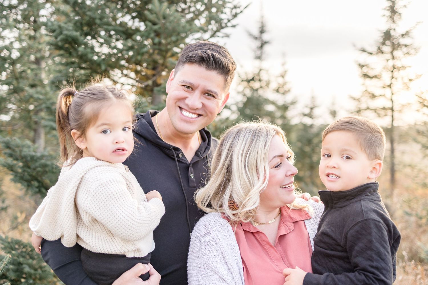 Natural outdoor family portrait session among evergreen trees at golden hour.
