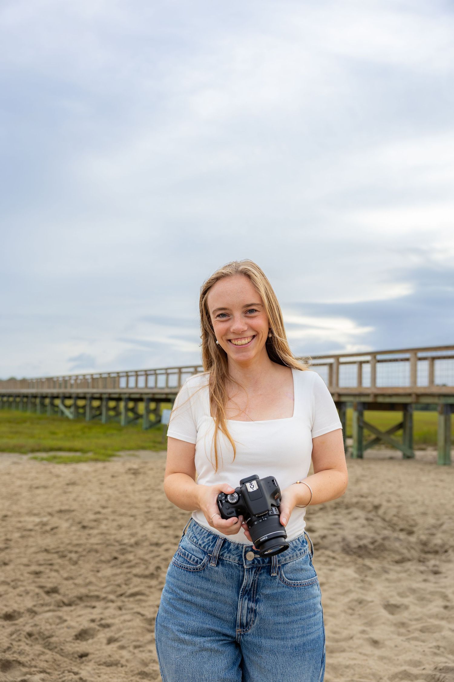 A photographer in jeans and white top stands on a sandy beach near a wooden pier on an overcast day.