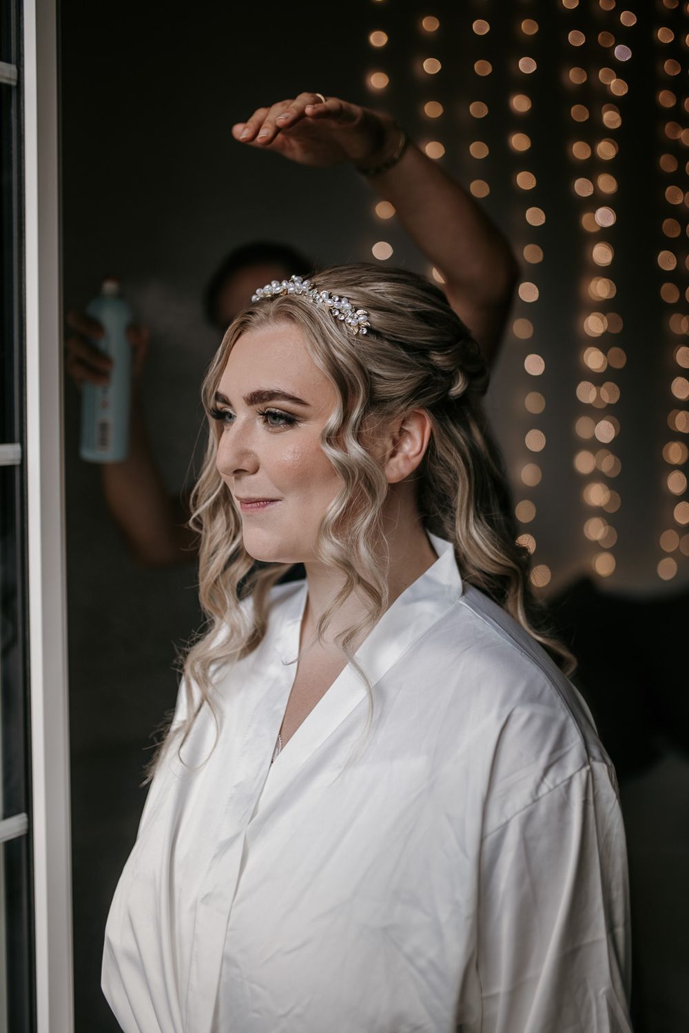 A blonde bride wearing a crystal hair crown gets her hair finished with hairspray against fairy lights.