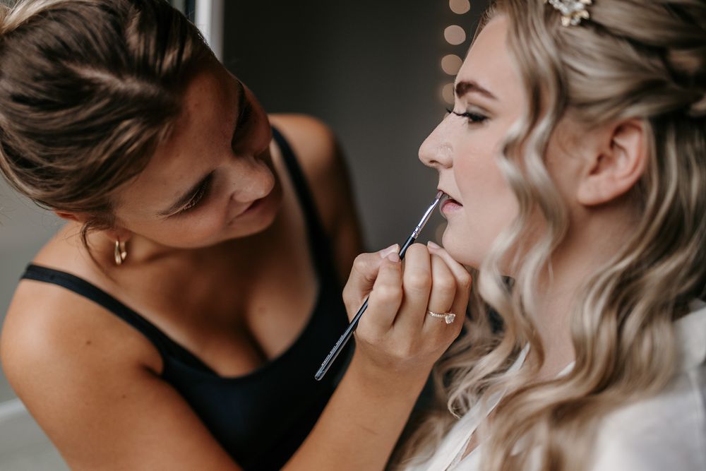 A makeup artist applies lipstick to a blonde bride with wavy hair and floral hair accessories.