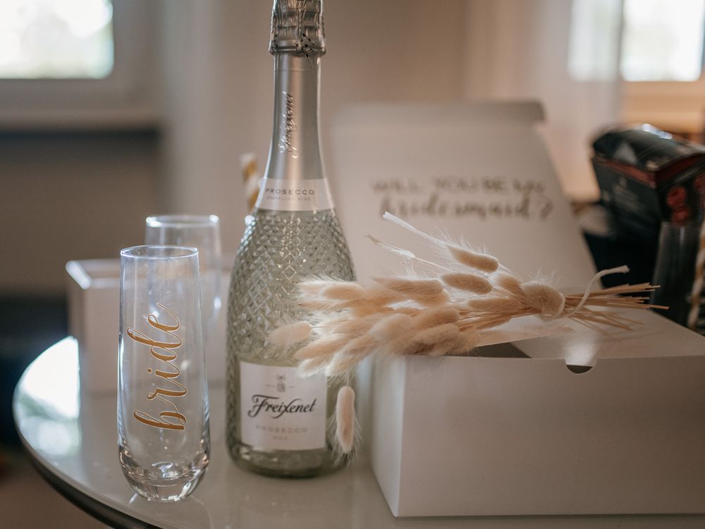 A Freixenet champagne bottle sits beside a bride glass and bridesmaid proposal box on a glass table.