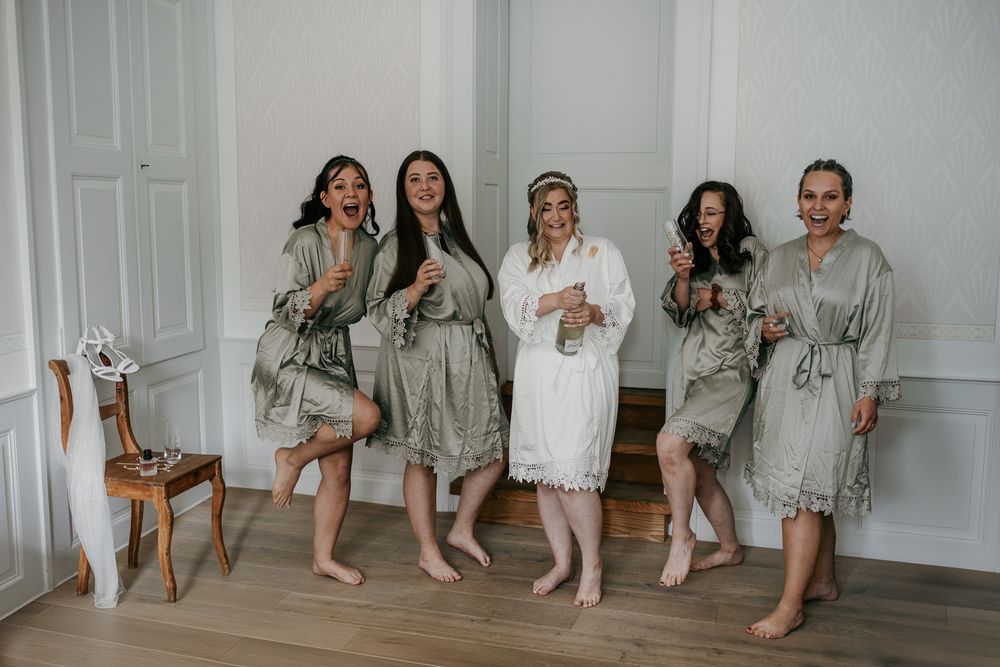 A bride in white lace robe celebrates with four bridesmaids in sage green robes holding champagne.