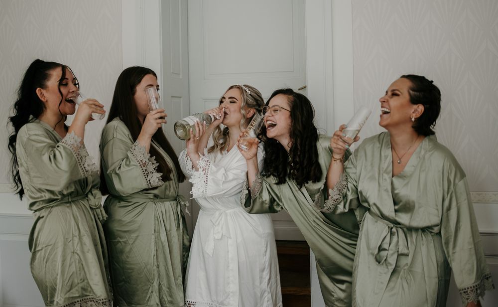 Bridesmaids in sage green satin robes celebrating with champagne alongside bride in white lace robe.
