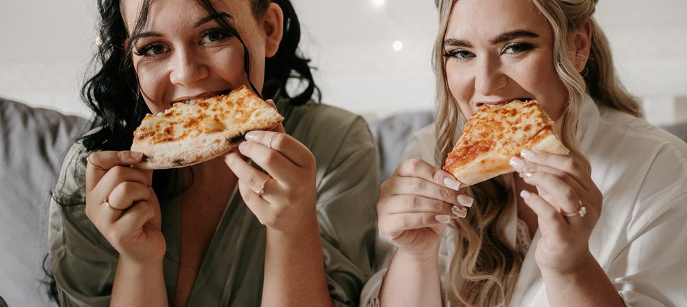 Two bridesmaids in satin robes smiling while eating cheesy pizza slices on wedding morning.
