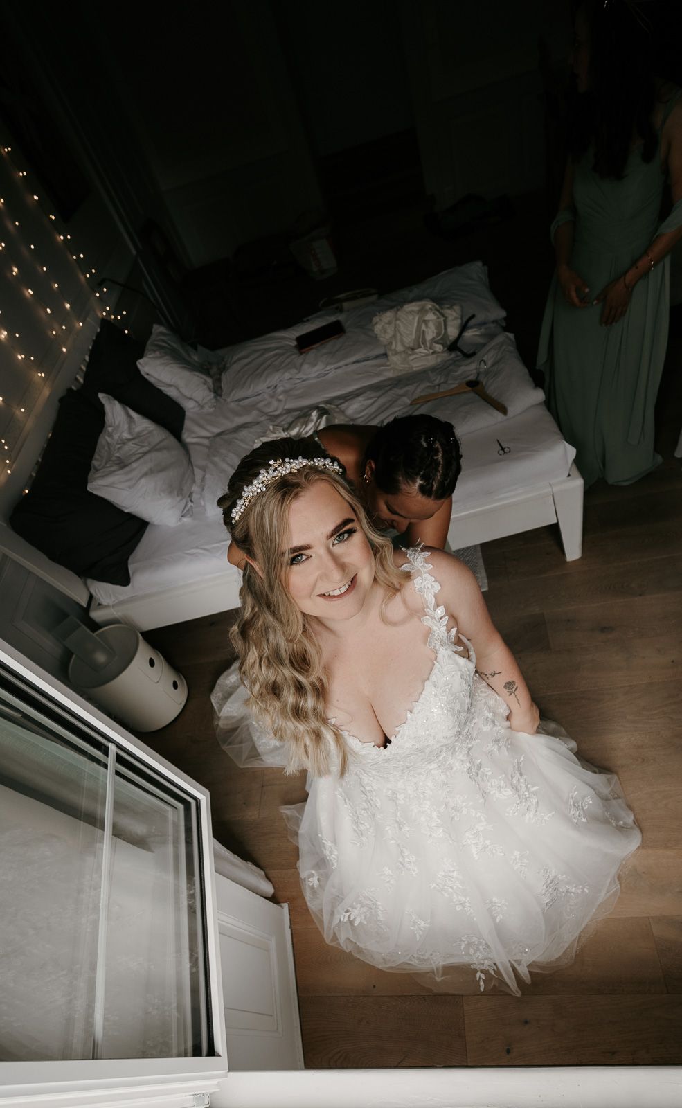 Blonde bride in floral lace wedding gown with crystal tiara smiling on staircase before ceremony.