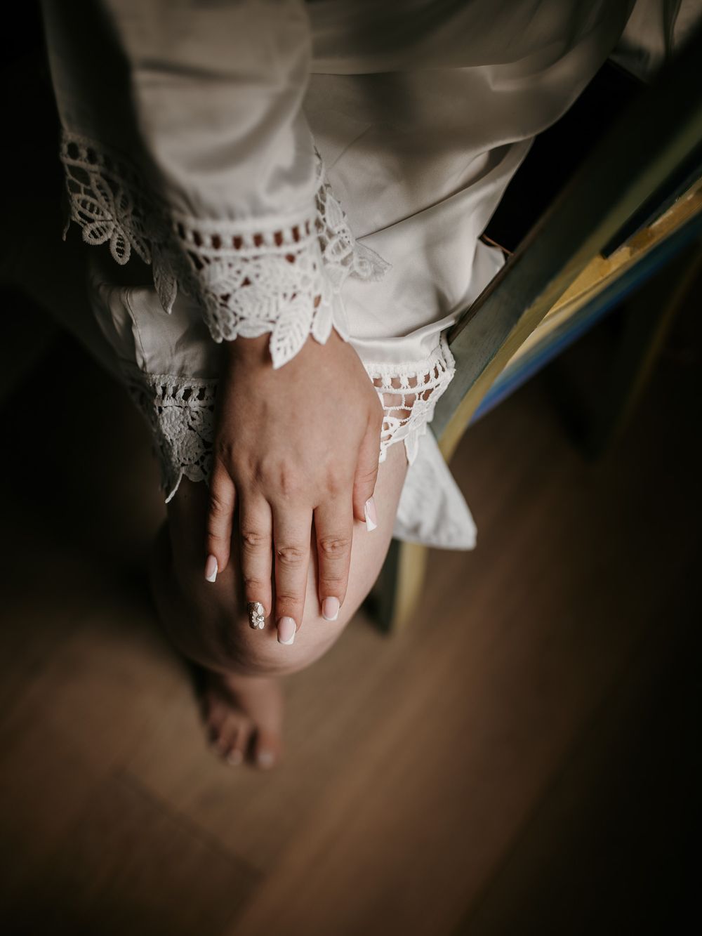 Close-up of bride's hands with French manicure resting on knees in delicate lace bridal robe.
