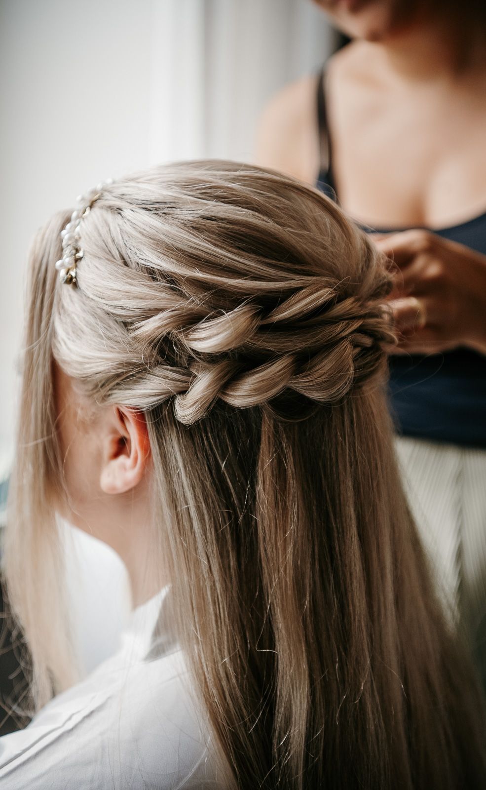 A hairstylist creates an elegant half-up crown braid with small floral pins in long blonde hair.