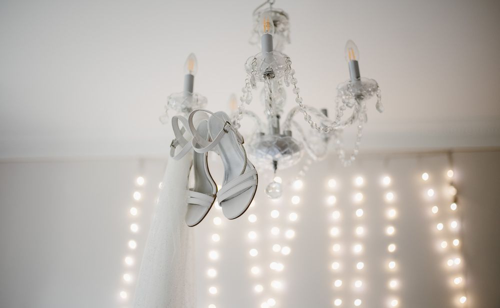 Silver bridal heels hang delicately from a white crystal chandelier against a backdrop of fairy lights.