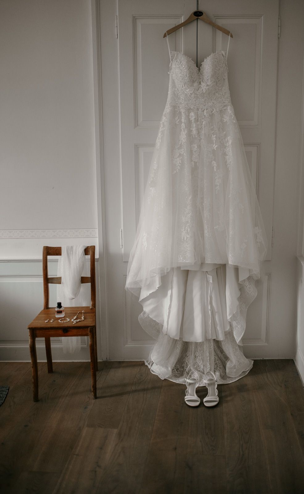 A white lace A-line wedding dress hangs on a wall beside a wooden chair with bridal accessories below.