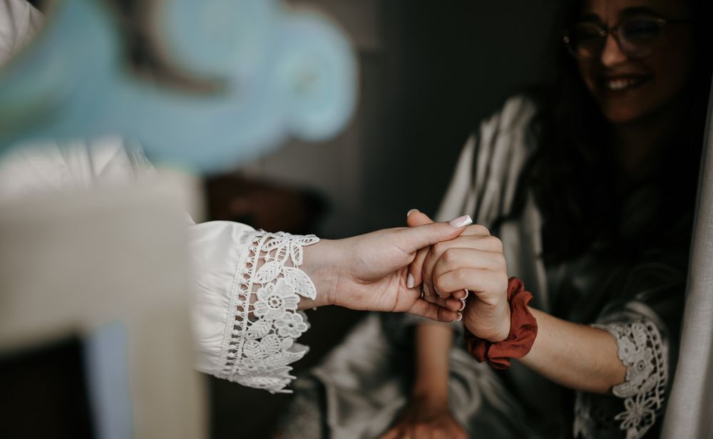 Two women in bridal robes share a warm handhold moment, one wearing lace cuffs and a red bracelet.