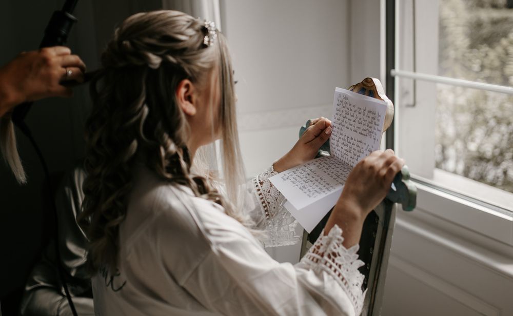 A bride in a white lace robe reads a heartfelt letter by a sunlit window while her hair is styled.