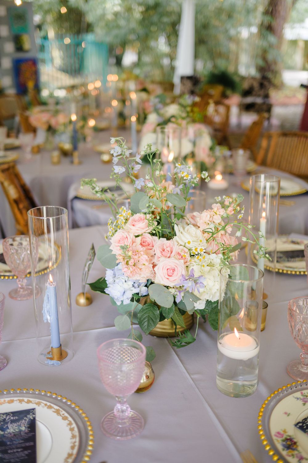 Elegant wedding table setting with pink and white floral centerpiece, gold-rimmed plates, and glowing candles on gray linens.