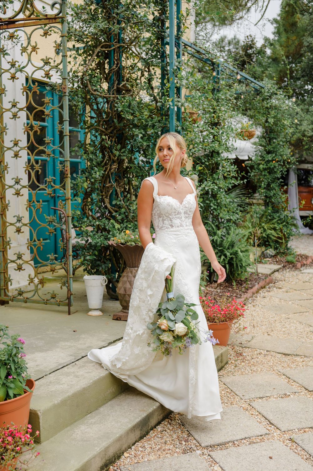 An elegant wedding gown flows down stone steps in a garden setting with ornate turquoise metalwork.