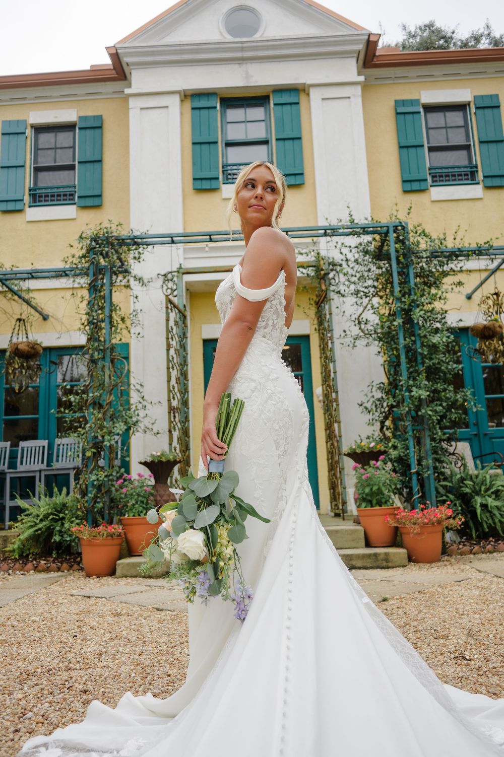 Elegant white wedding gown photographed against a yellow colonial-style house with teal shutters and climbing vines.