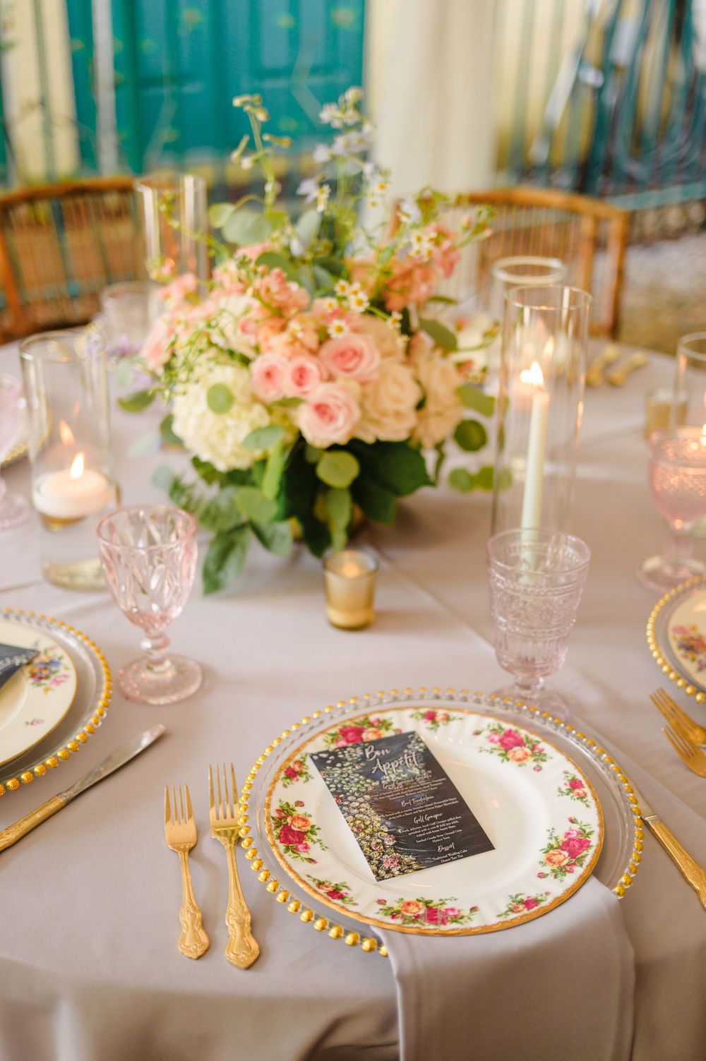 Elegant wedding table setting with floral china plates, gold cutlery, and a pink and white rose centerpiece.