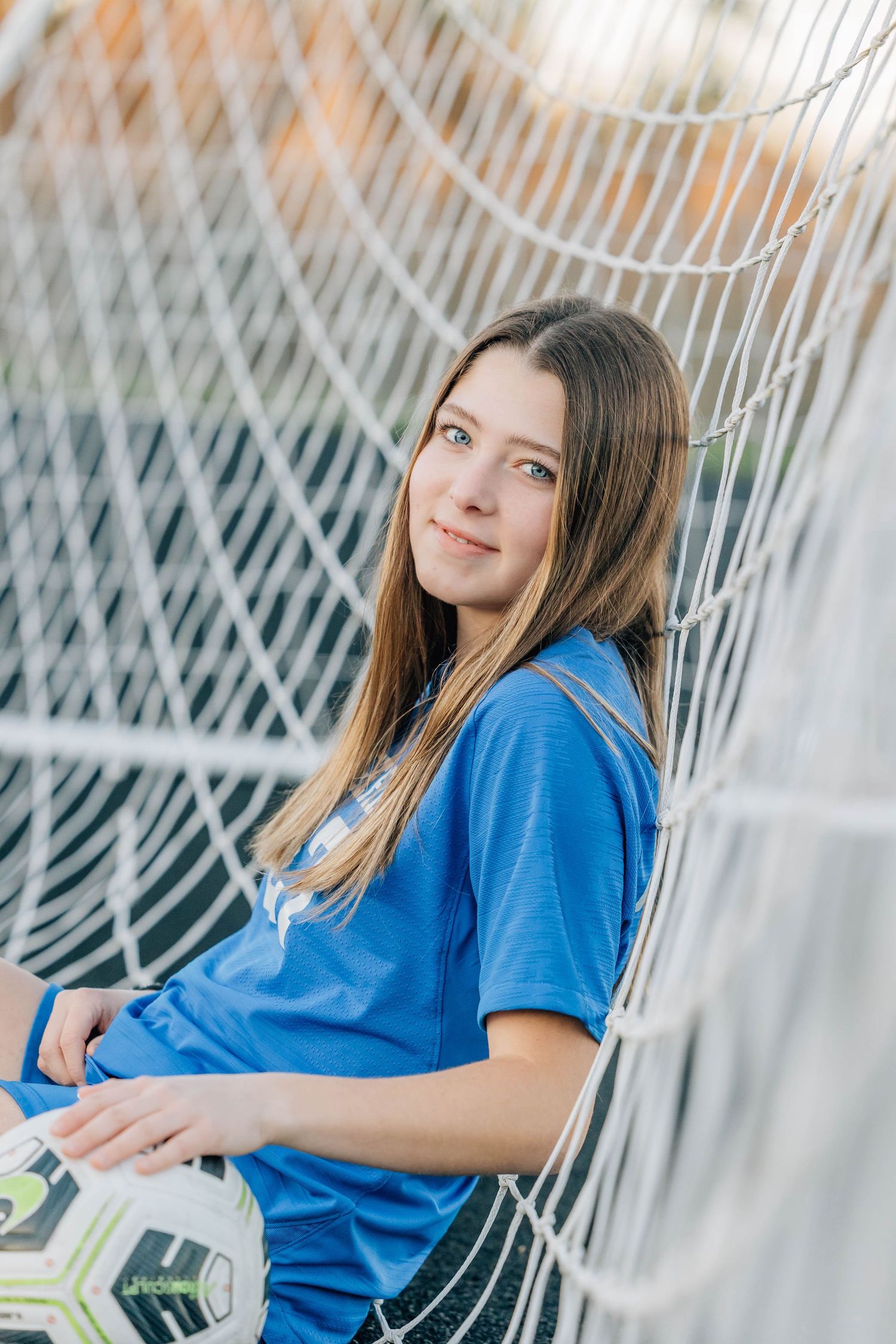Teen girl in blue soccer jersey leaning against goal net holding soccer ball.