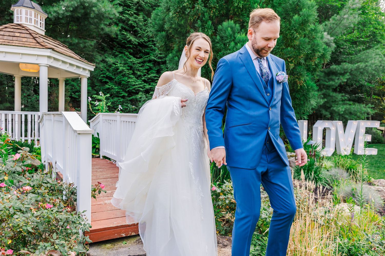 A couple in wedding attire walks through a lush garden path with the bride wearing white and groom in a blue suit.