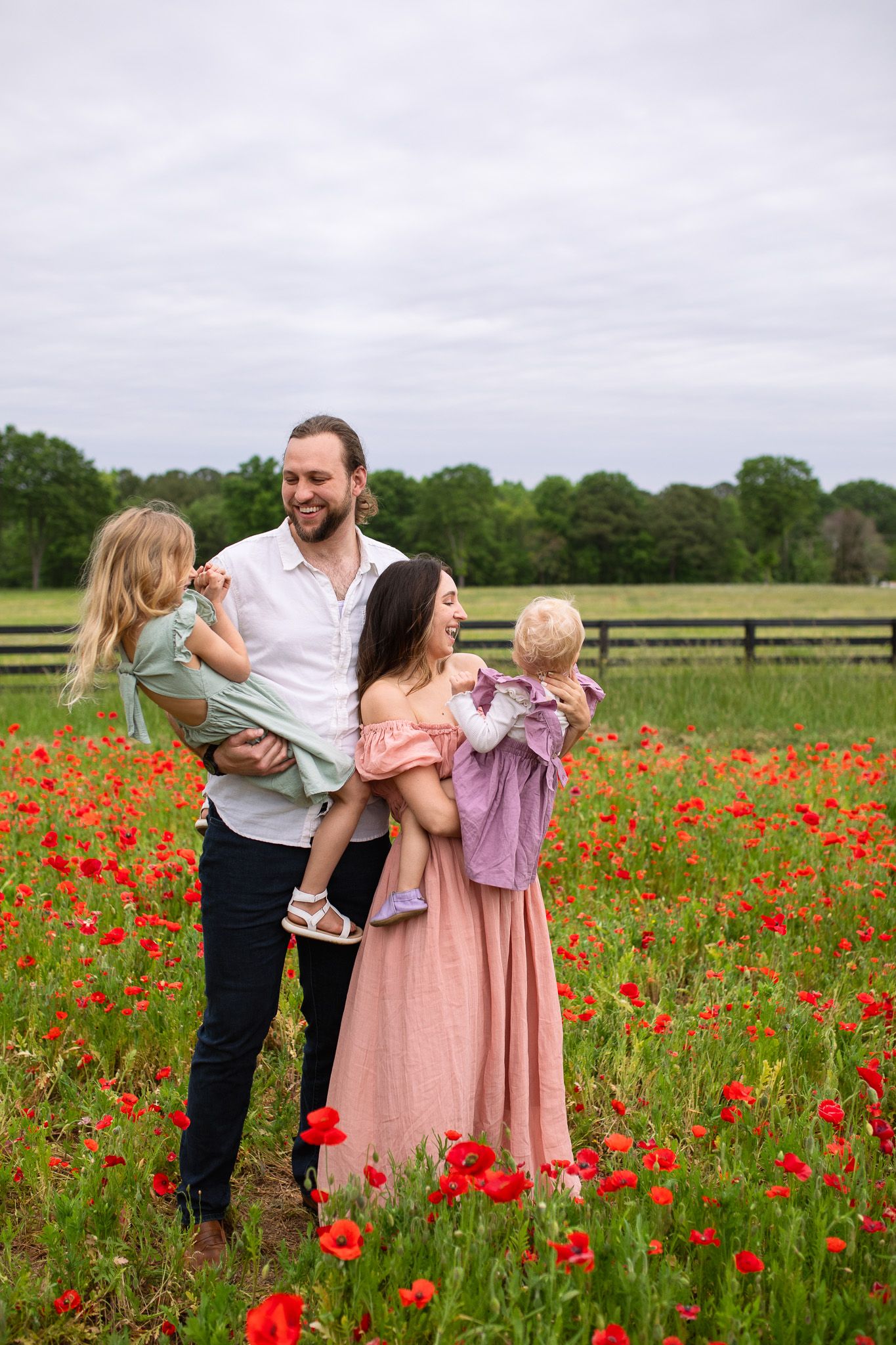 Family standing together in a vibrant red poppy field with wooden fences and green trees in the background.