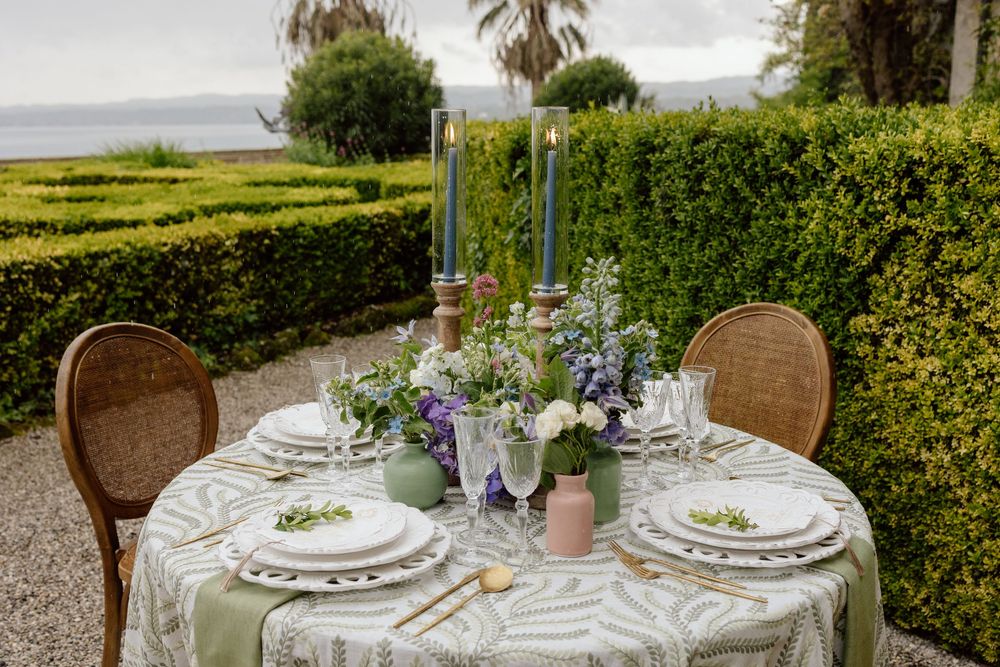 Elegant outdoor dining table with wicker chairs set against manicured hedges and ocean view, featuring floral centerpieces.