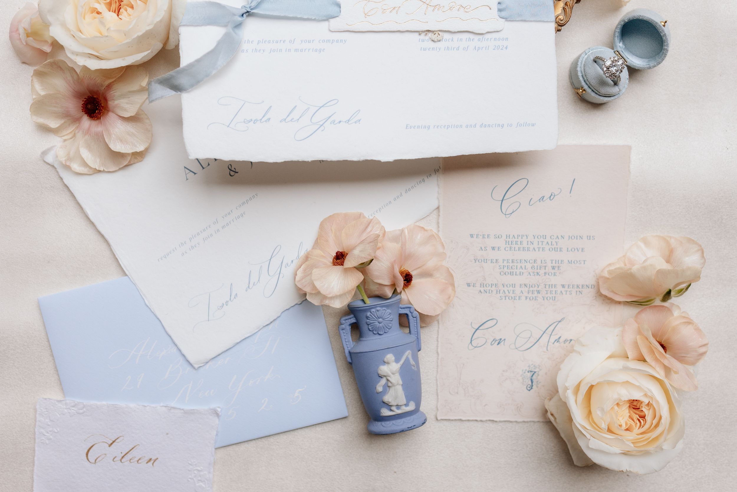Elegant wedding stationery flat lay with calligraphy invitations, peach roses, and dusty blue accents on white surface.
