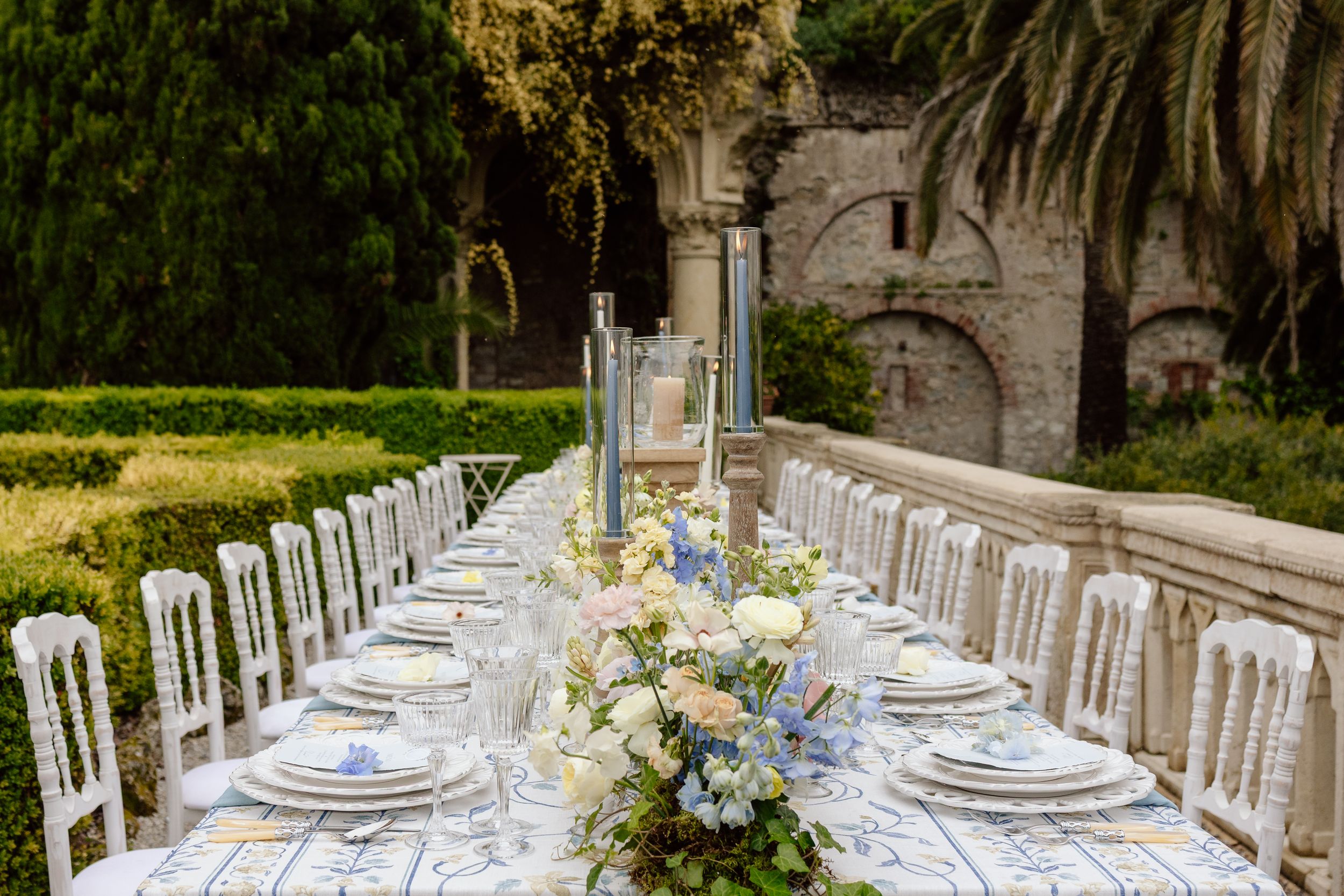 Elegant outdoor wedding reception table setting with white chairs and delicate floral centerpieces extends through a garden path.