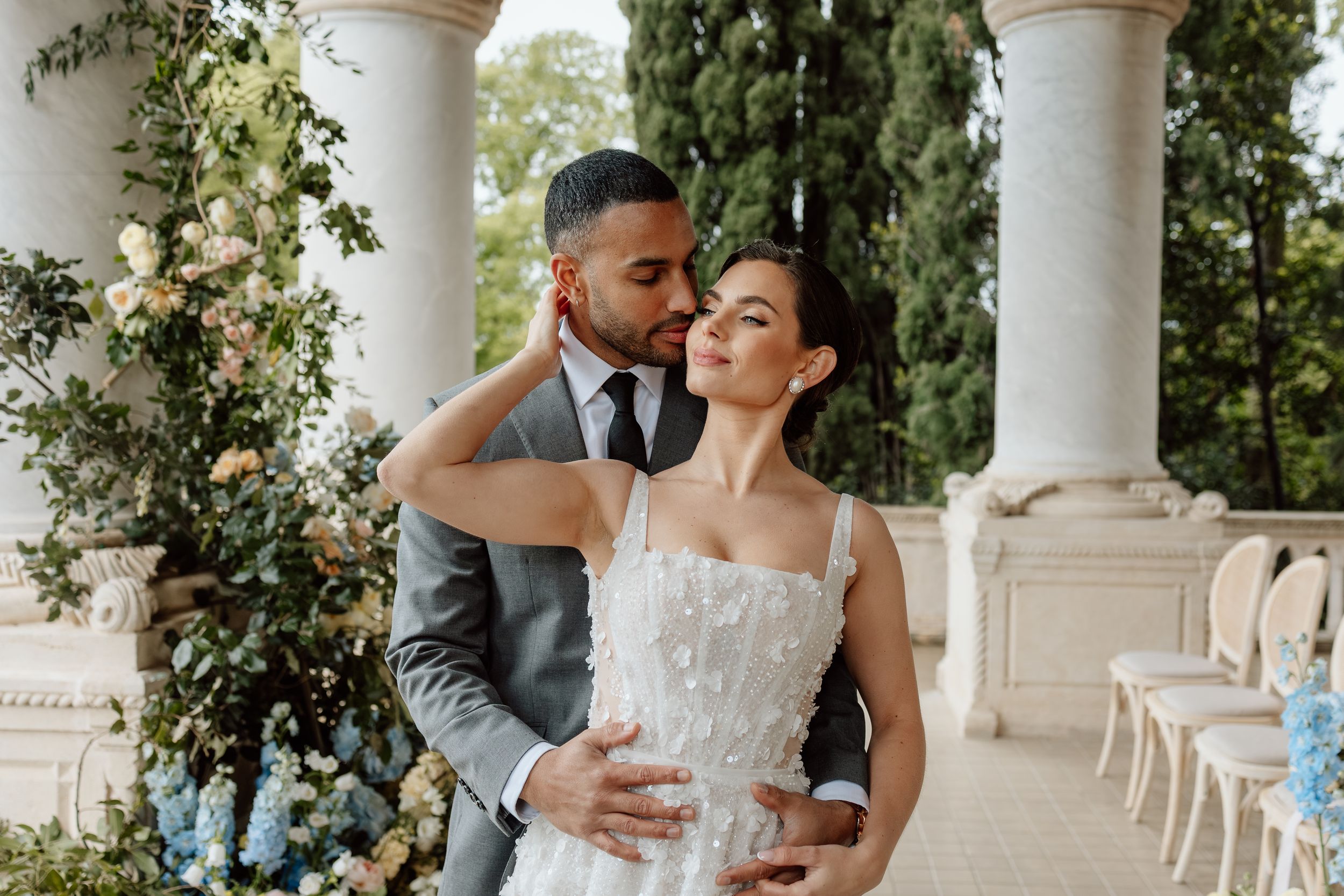 A tender romantic moment between a couple in wedding attire at an elegant venue with white columns and floral decor.