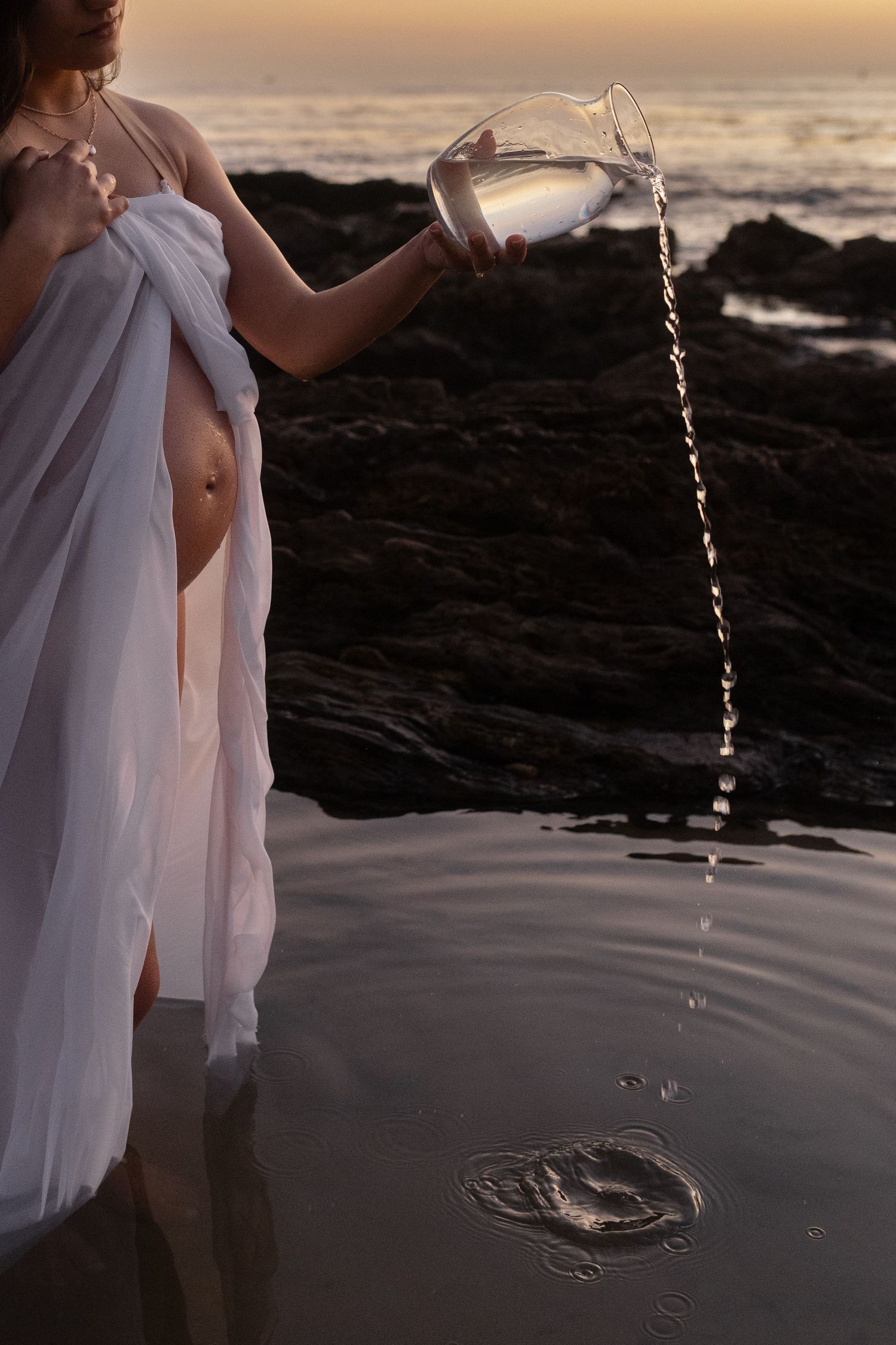 Water droplets falling into ocean at sunset with figures in white silhouetted against sky.