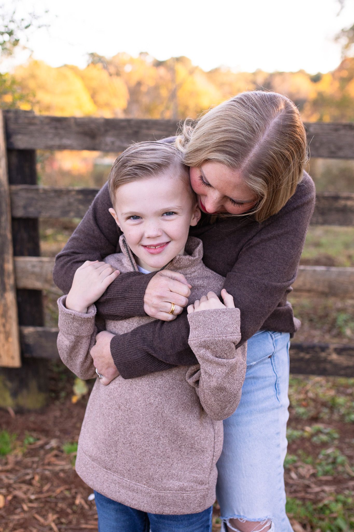 Loving mom and child share a warm embrace outdoors on an autumn day with rustic wooden fence in background at Sugg Farm