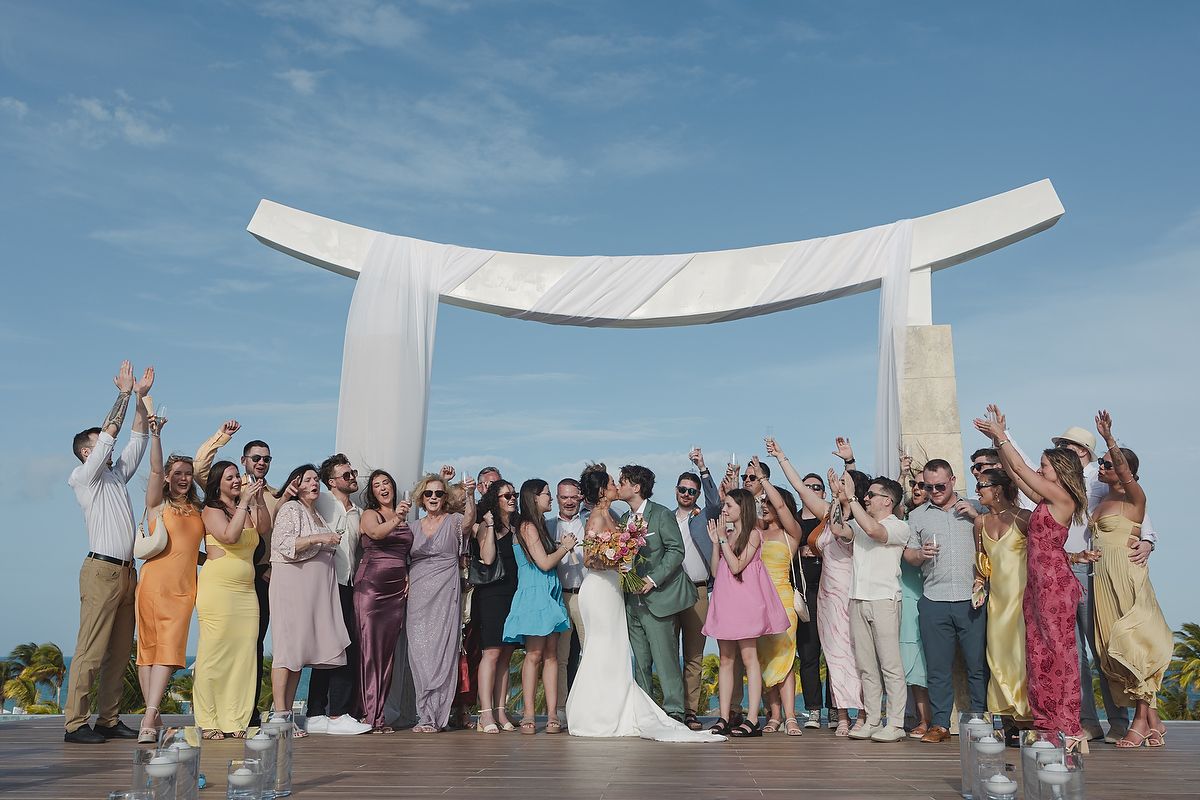 Wedding party celebrates under a white arch on a sunny beach, with guests in colorful attire raising their arms in joy.
