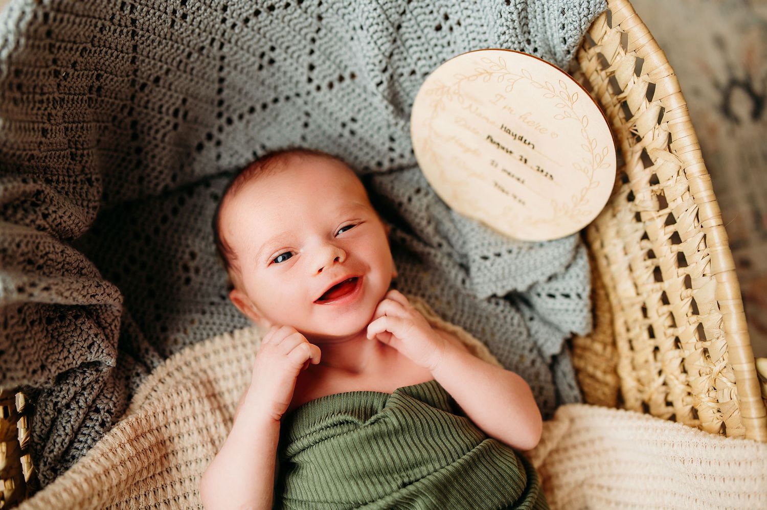 A smiling baby laying in a woven basket wrapped in soft neutral fabrics in Rochester MN