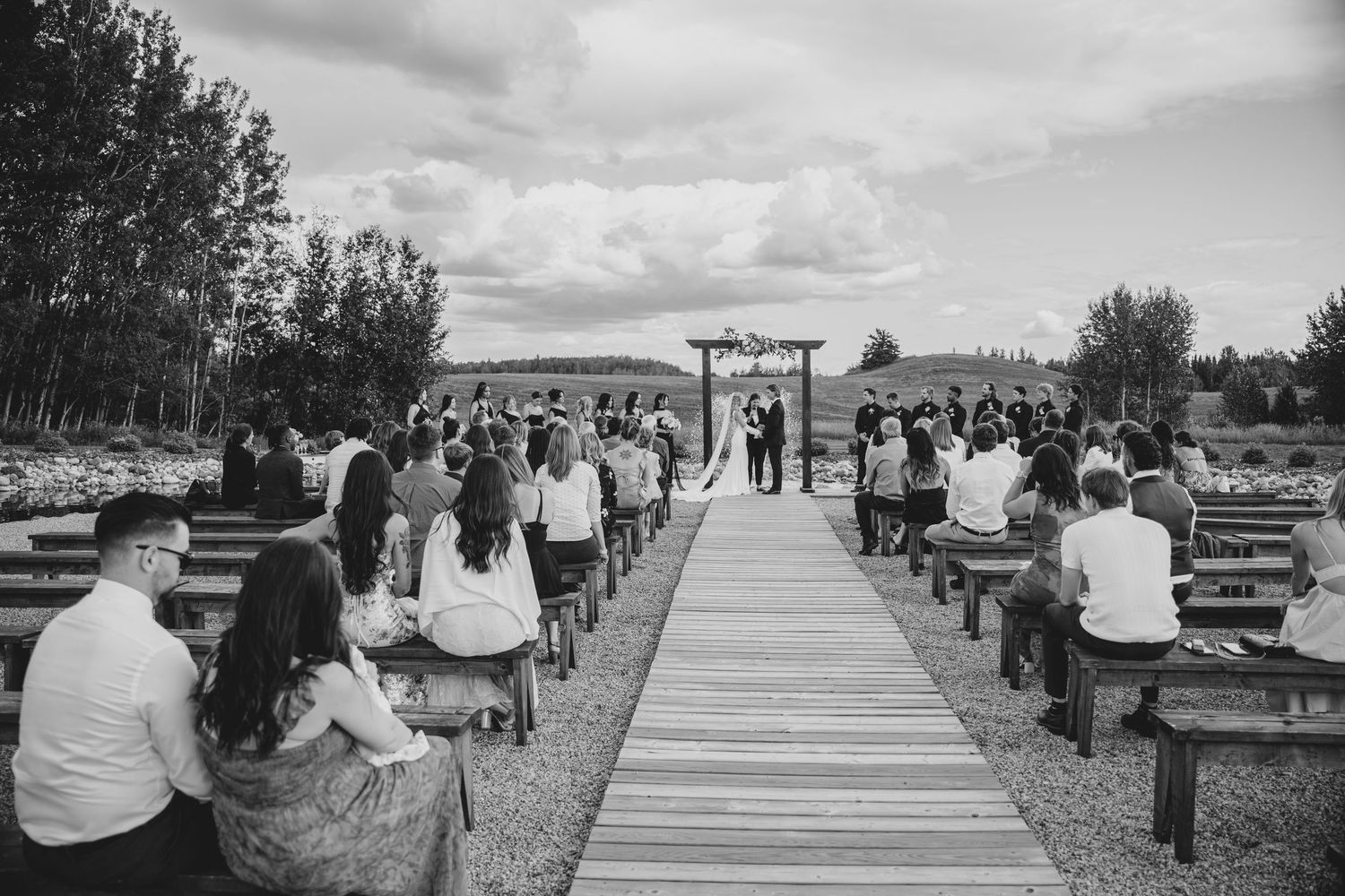 Guests gather for an outdoor wedding ceremony on a wooden boardwalk path surrounded by trees and cloudy skies.