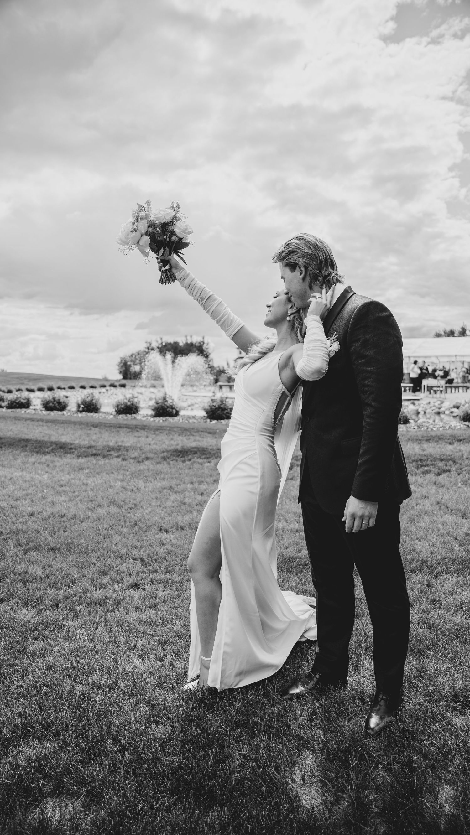 A black and white wedding photo of a couple in formal attire celebrating outdoors with the bride raising her bouquet.