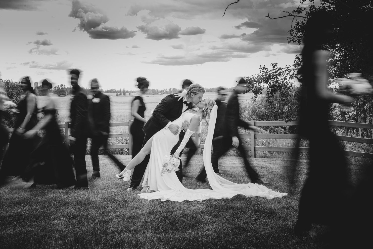Black and white artistic wedding photo showing a couple embracing outdoors with moving guests creating motion blur effect.