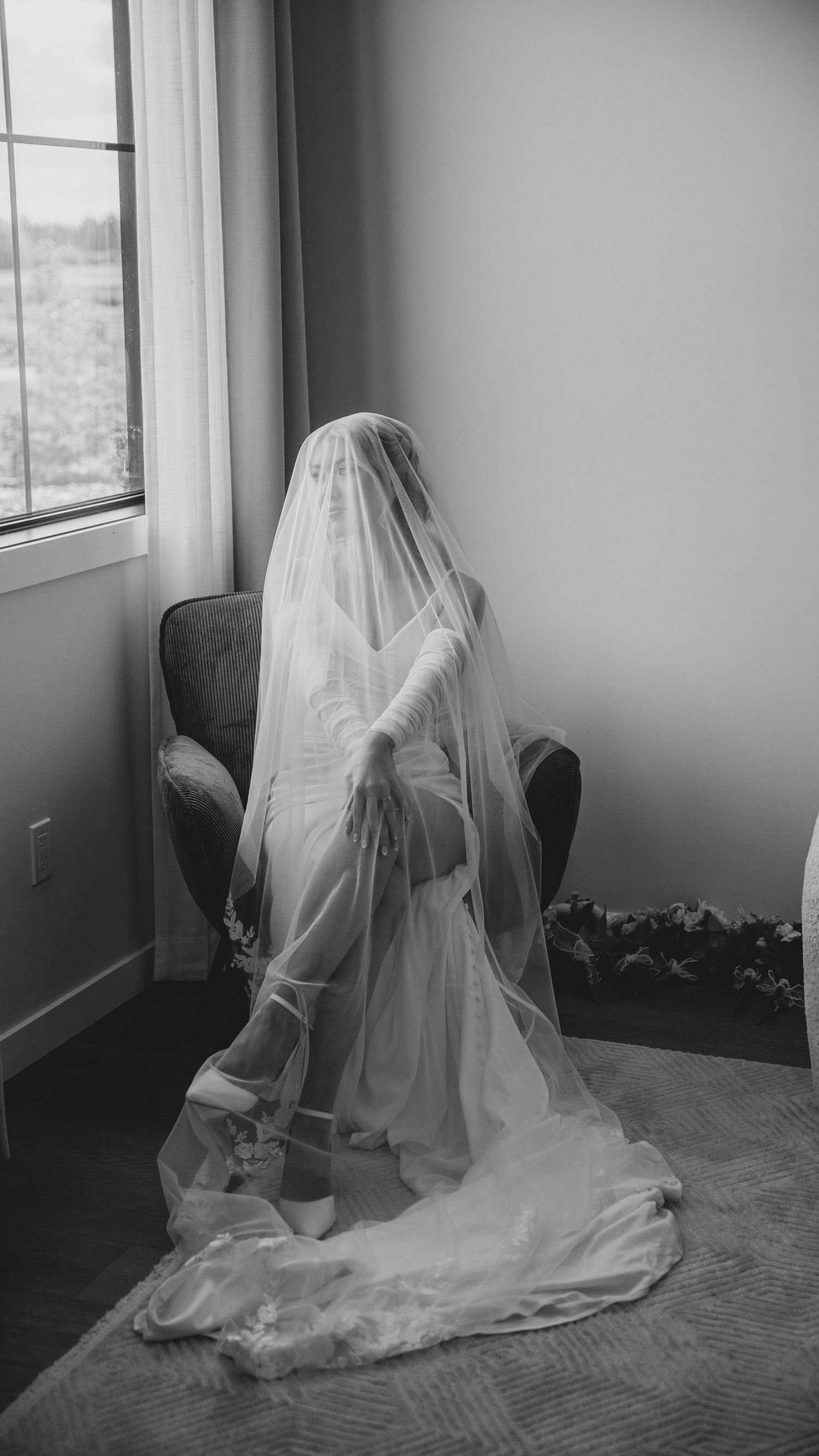 A figure in a flowing white wedding veil and dress sits contemplatively by a window in black and white.